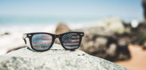 sunglasses with american flag on the lenses sitting on a rock with the beach in the backdrop